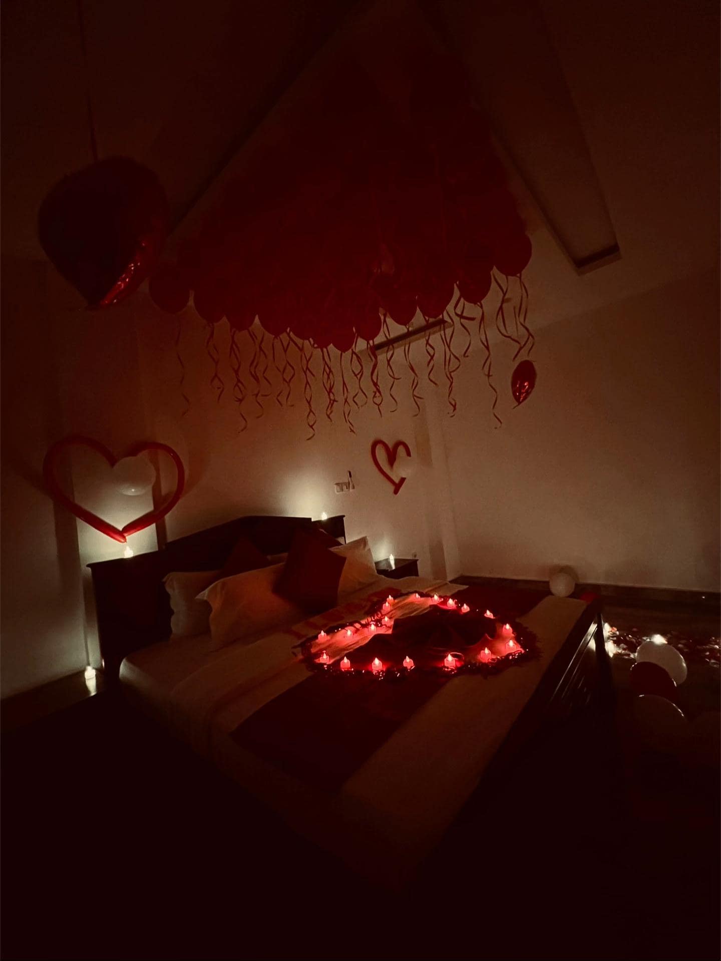 Chambre dressée pour la Saint-Valentin — cœurs en ballons au mur, bougies LED alignées et voûte de ballons rouges au plafond